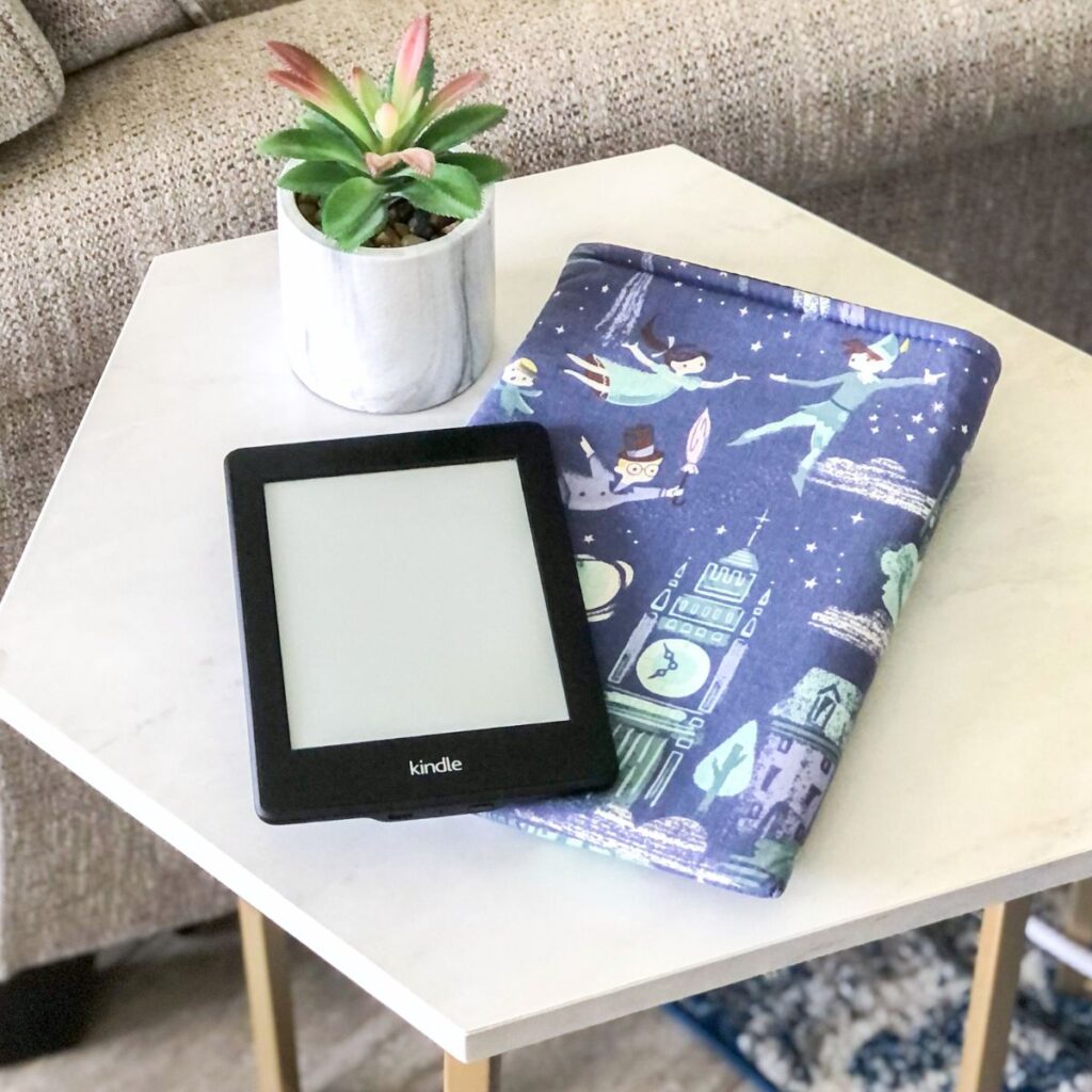 printed fabric book sleeve from a bookish merchandise shop next to an e-reader and a small houseplant