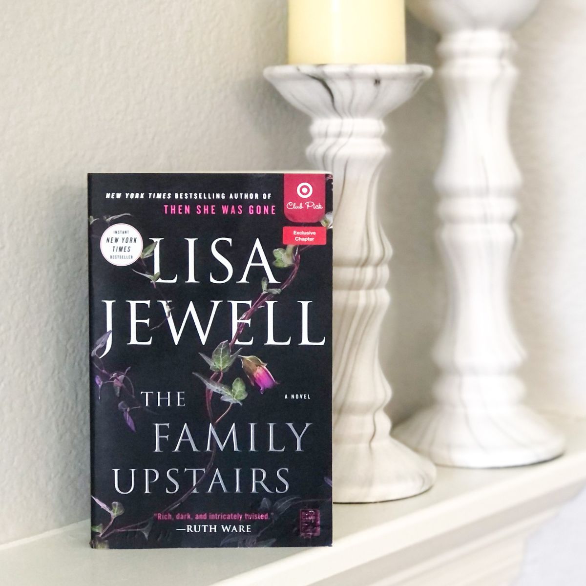 The Family Upstairs book by Lisa Jewell sitting on a fireplace mantel with white candle sticks behind it