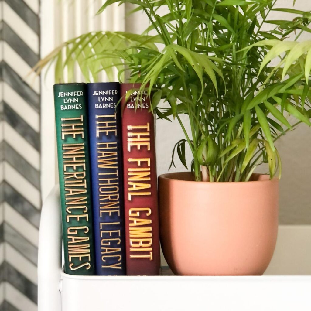 The Inheritance Games books stacked on a white book cart with a house plant