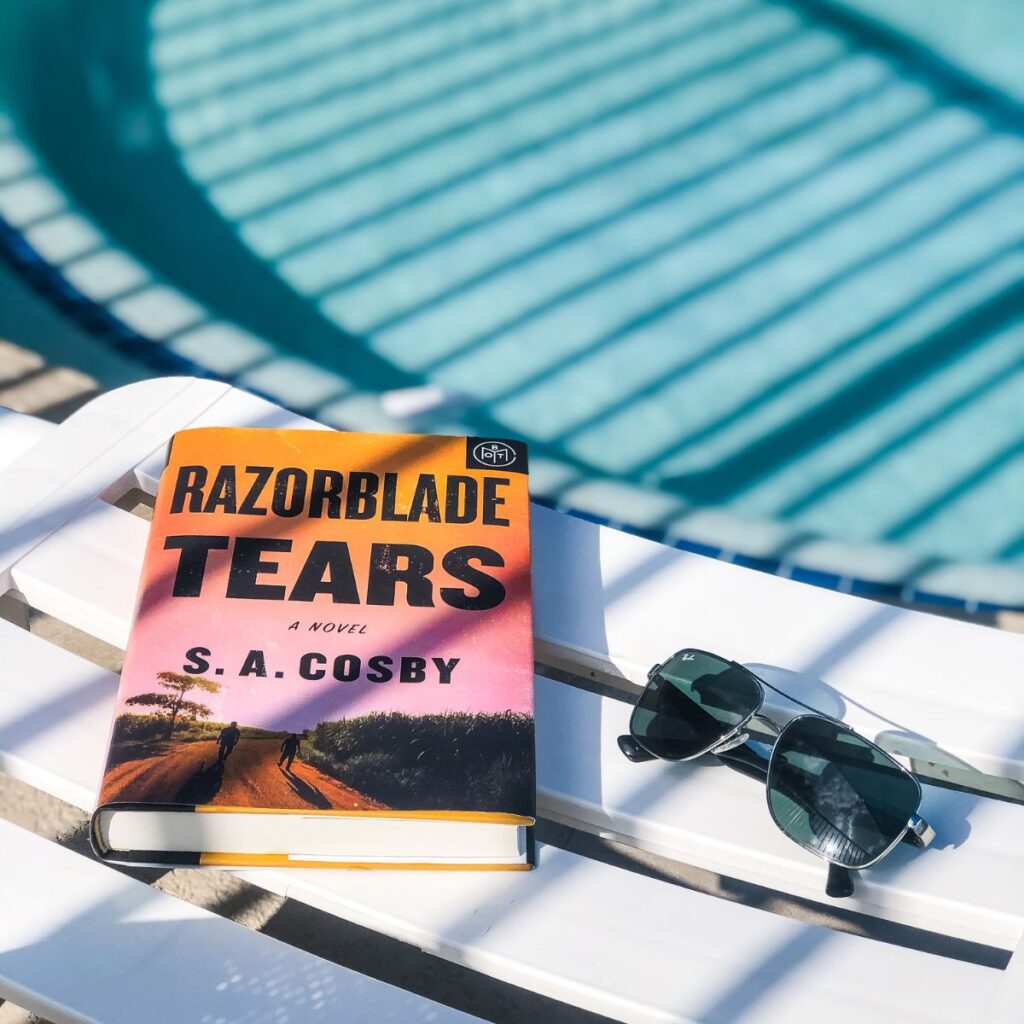 a hardcover book and pair of sunglasses sitting on a white lounge chair next to a pool