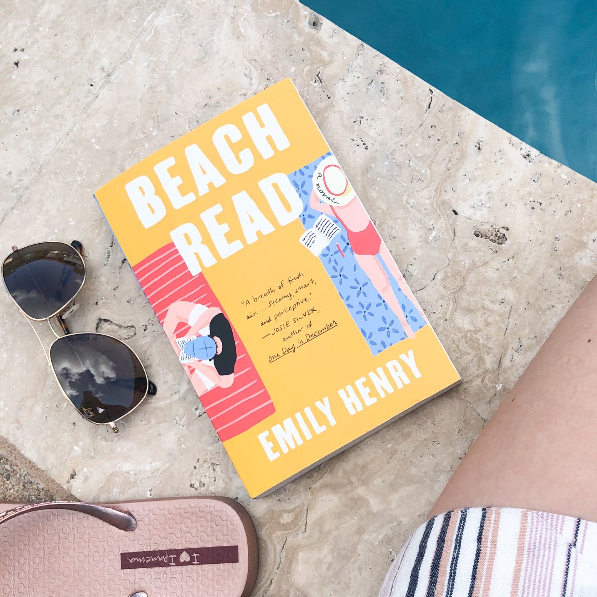 Beach Read paperback by Emily Henry sitting by a pool with sunglasses and flip flops beside it