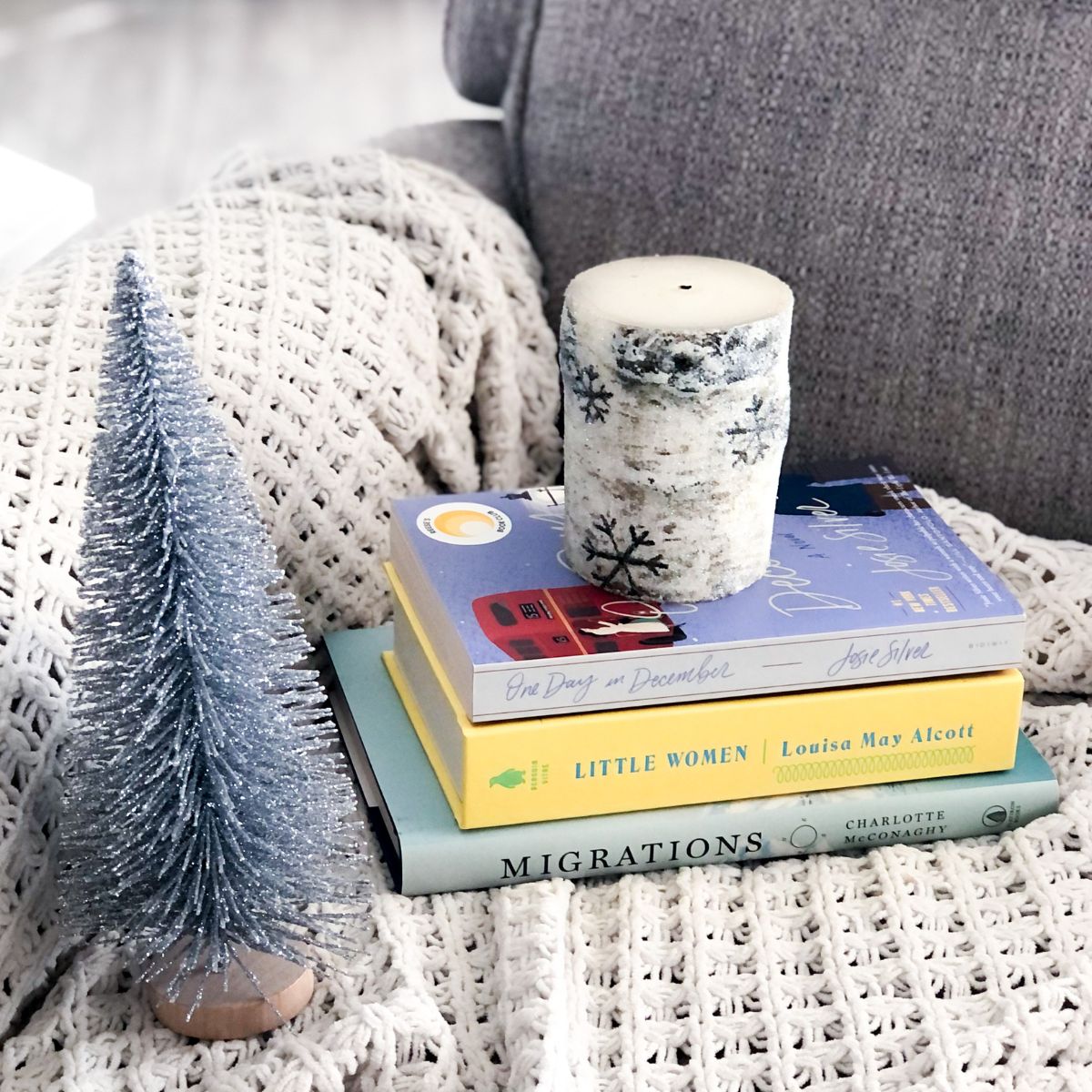 stack of winter books with a candle on top and and a mini silver tree next to it on a grey chair