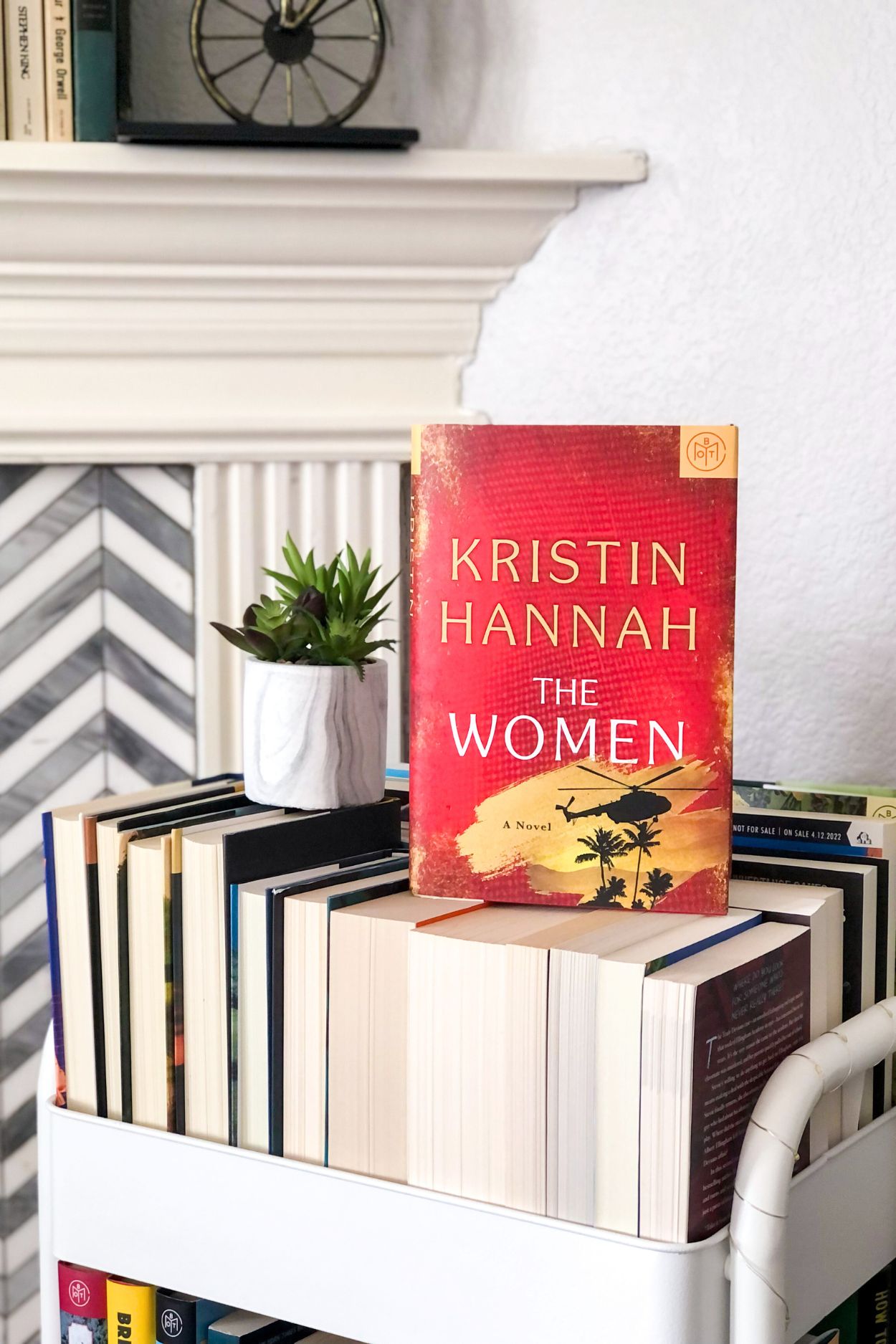 The Women by Kristin Hannah hardcover book sitting on a book cart on top of a pile of paperback books