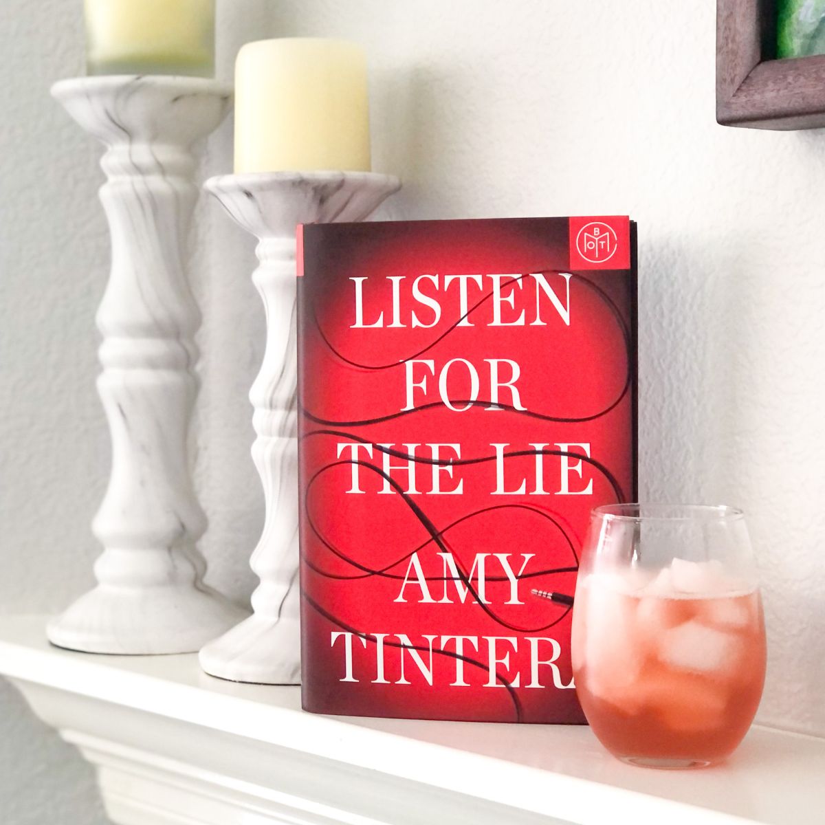 Hardcover Listen for the Lie book on a white fireplace mantel next to a stemless wine glass filled with pink kombucha and white marble candle holders