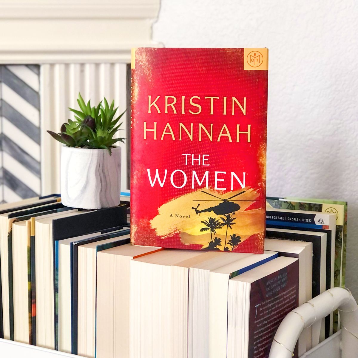 The Women hardcover book sitting on a book cart with a small plant beside it.